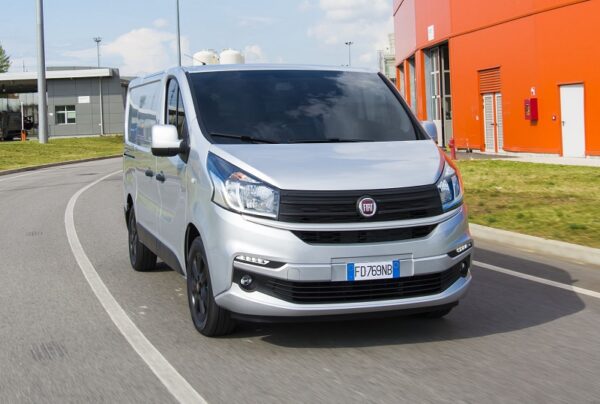 White Fiat Talento van driving on road with orange industrial building in background.