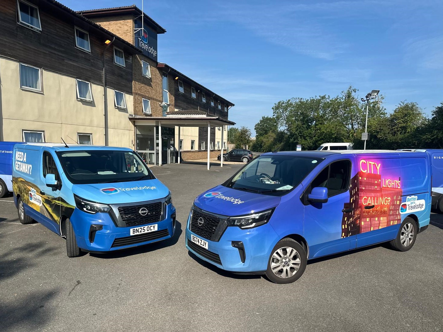Two bright blue Nissan vans parked outside a Travelodge hotel building, one displaying City Cleaning branding.