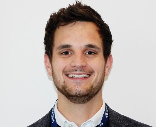 Man with dark hair and beard smiling at camera, wearing dark cardigan and white shirt with blue lanyard.