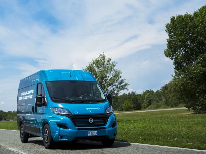 Bright blue electric van parked on rural road with green fields and trees under cloudy sky.