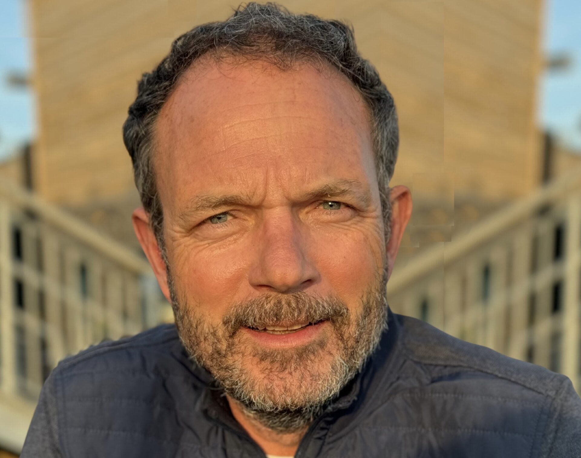 Adrian Simpson, man with grey beard wearing dark shirt, portrait against architectural background.