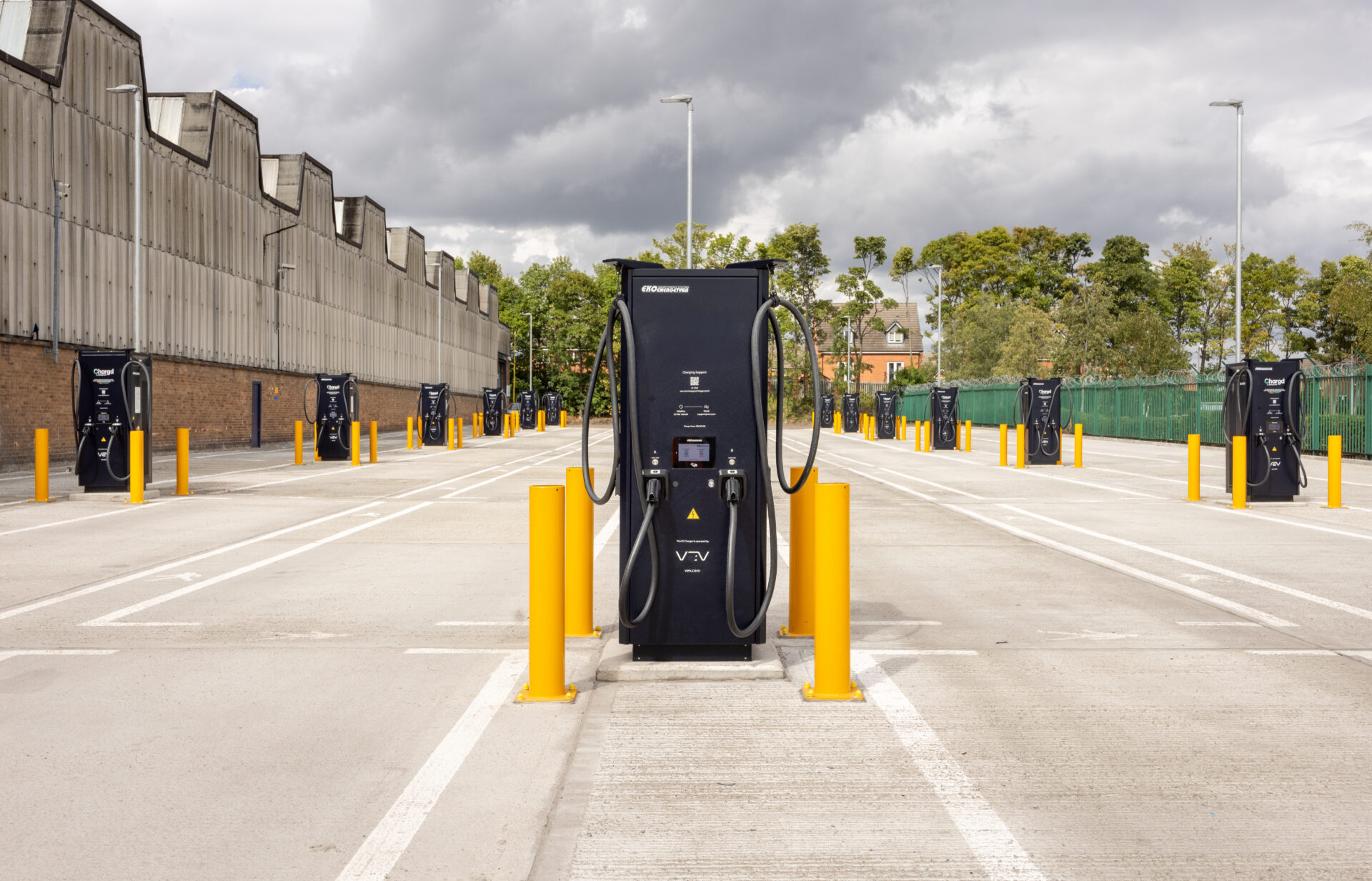 Row of electric vehicle charging stations with black chargers flanked by yellow bollards on an outdoor paved forecourt,…