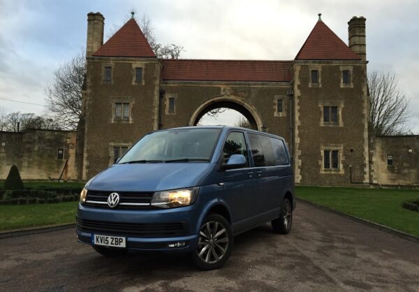 Blue Volkswagen Transporter Kombi van parked in front of a stone gatehouse with twin towers and archway.