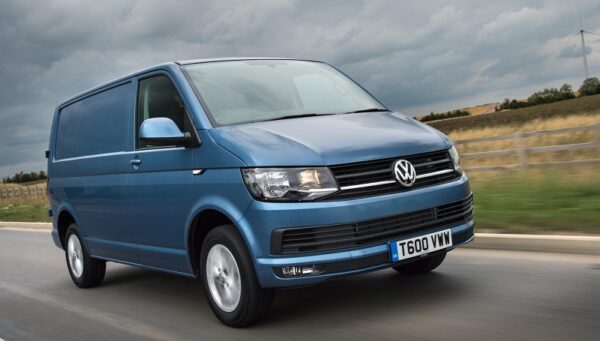 Blue Volkswagen Transporter T6 van driving on a road with fields visible in the background.