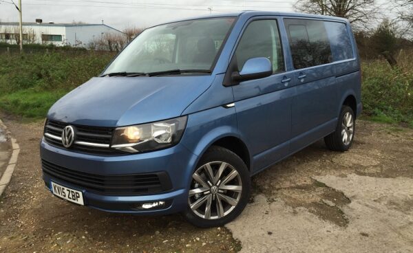 Blue Volkswagen Transporter Kombi van parked on gravel drive, headlights on, bare trees in background.