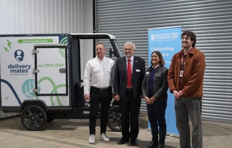 Five people stand beside a small black electric delivery vehicle branded with Delivery Mates logo at an industrial…