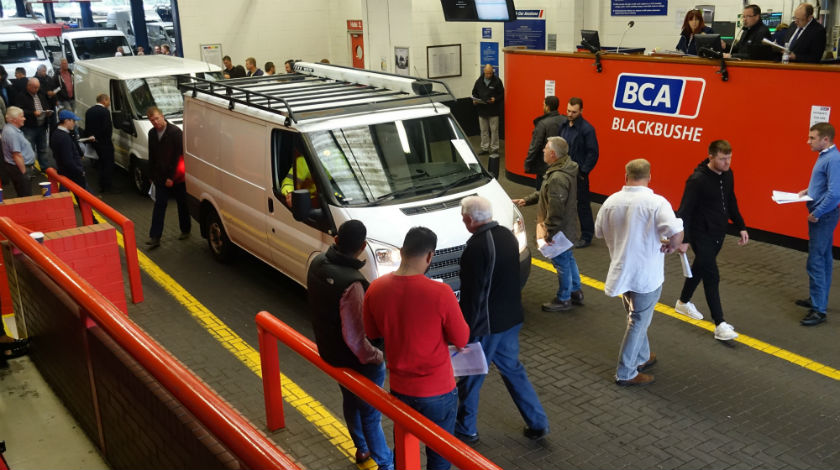 Visitors examine a light commercial vehicle on display at a BCA Blackbushe auction hall with staff and red barriers.