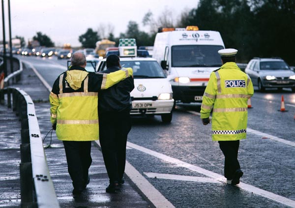 Traffic officers in high-visibility jackets converse at incident scene on wet motorway with emergency vehicles behind.