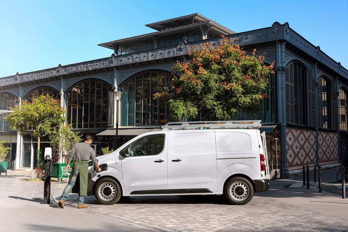 White electric van parked outside Victorian market hall, driver consulting clipboard with historic building and autumn…