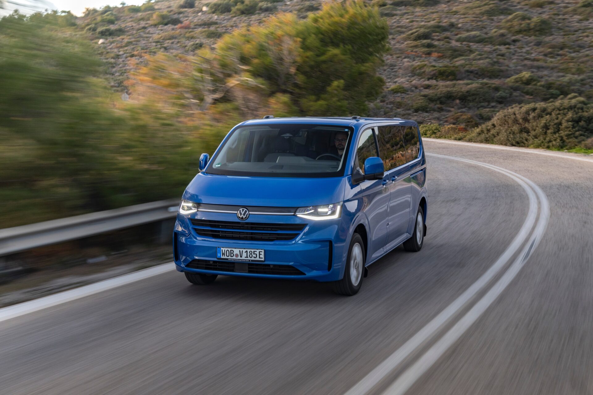Blue Volkswagen e-Transporter van driving on a curved mountain road with scrubland hillside in background.