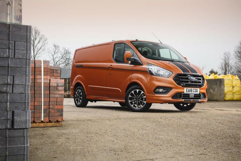 Orange Ford Transit Custom van parked beside stacked building materials and pallets on industrial site.