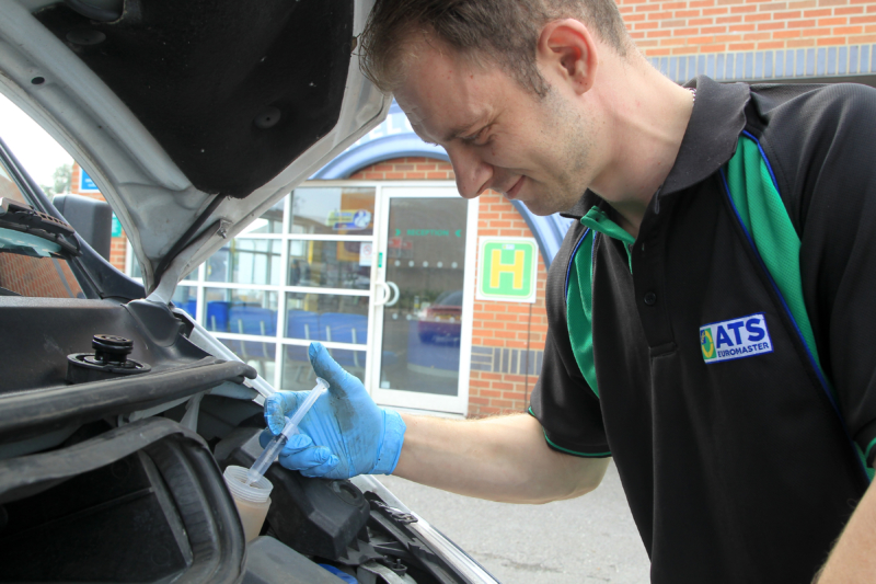 Mechanic in black and green uniform wearing blue gloves inspecting under car bonnet during winter maintenance check.