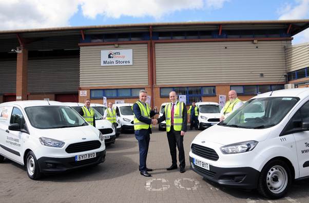 Five workers in high-visibility jackets stand between white Ford vans parked outside an industrial warehouse building…