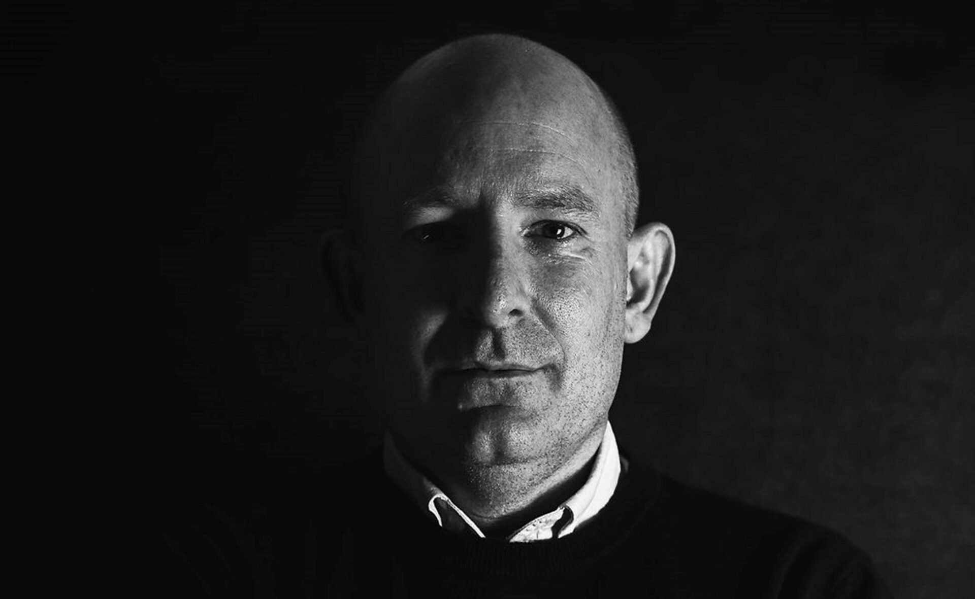 Bald man in dark jumper with white collar against black background, black and white portrait photograph.
