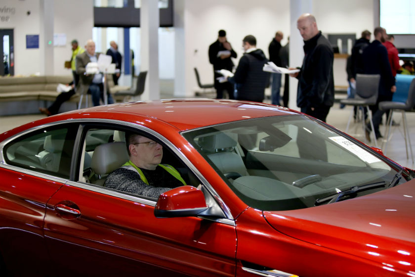 Red car on display with man in driver's seat at indoor event with attendees in background.
