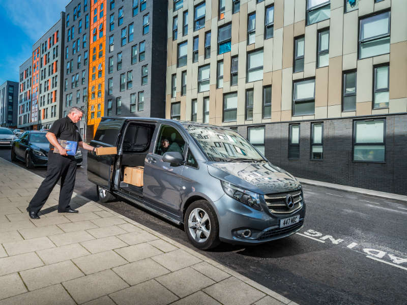 Delivery driver loading parcels into silver Mercedes van on urban street with apartment buildings behind.