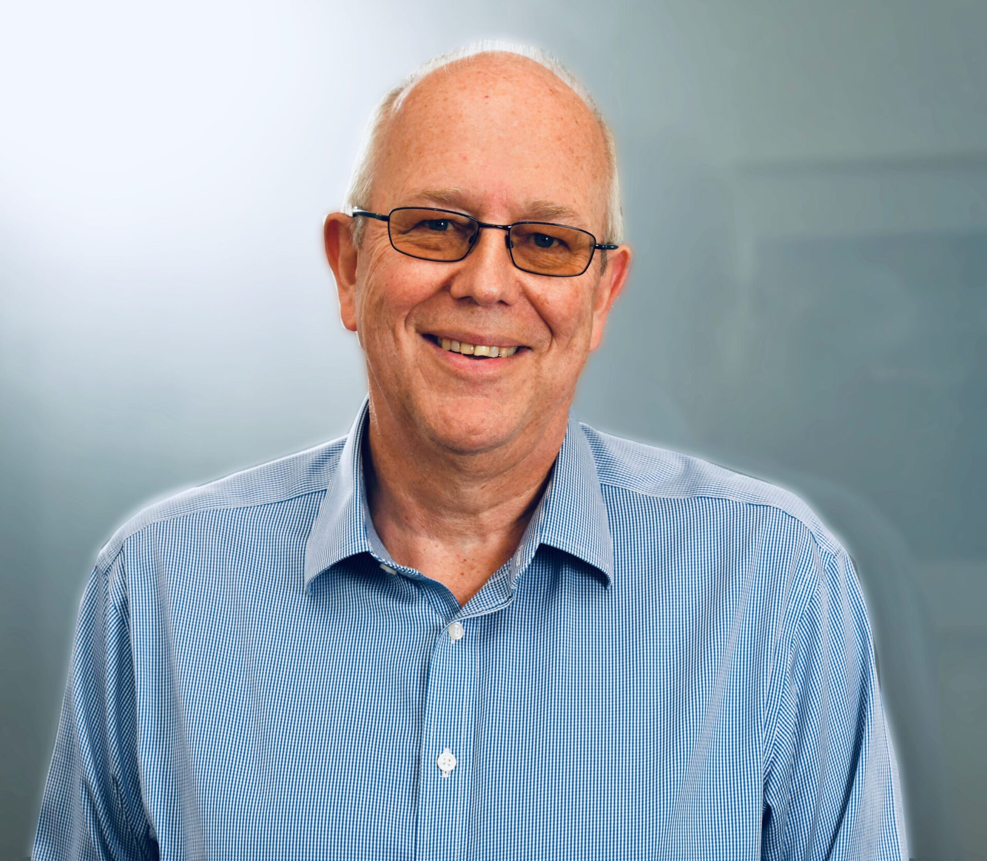 Bald man with glasses wearing light blue shirt, smiling at camera against pale blue background.