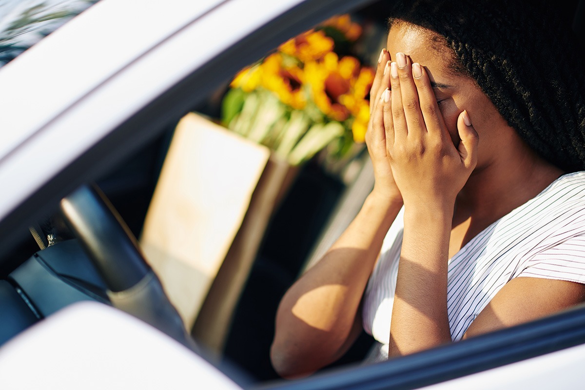 Woman sitting in car with hands to face in distress, with flowers visible in background.