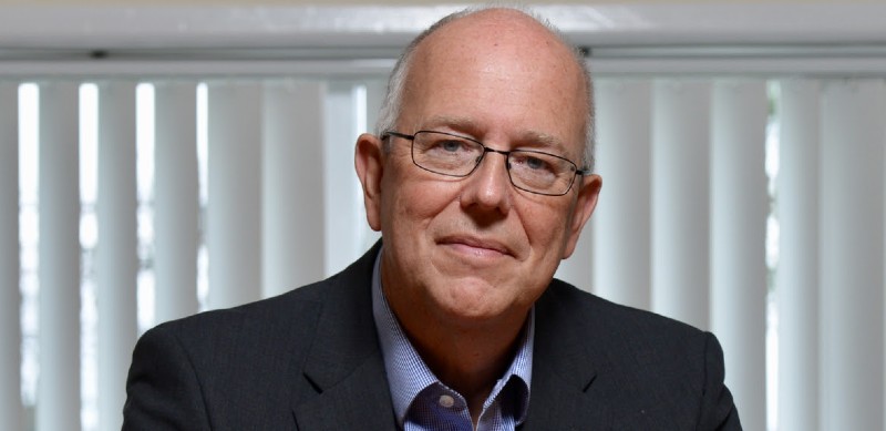 Bald man wearing glasses and dark blazer over blue shirt, seated indoors with vertical blinds behind.