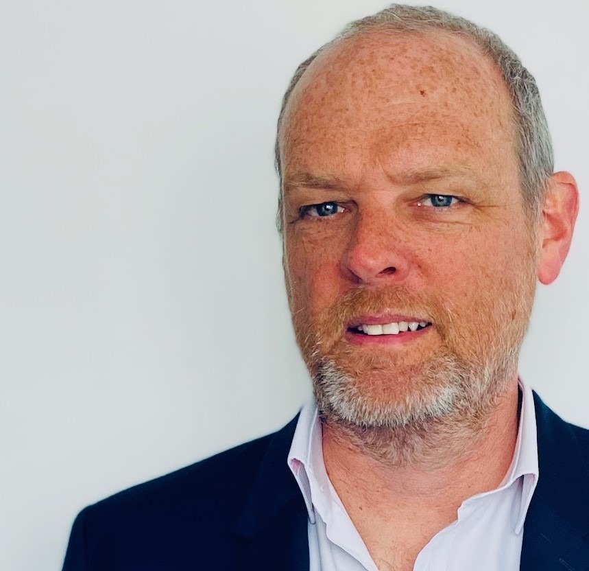 Middle-aged man with grey hair and beard wearing navy blazer and white shirt against white background.