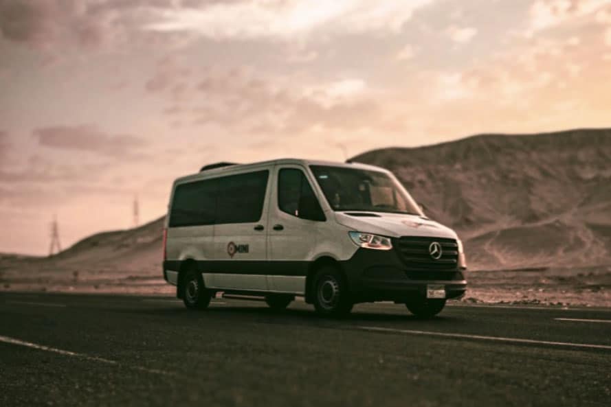 White Mercedes-Benz van parked on desert road with rocky hills in background.