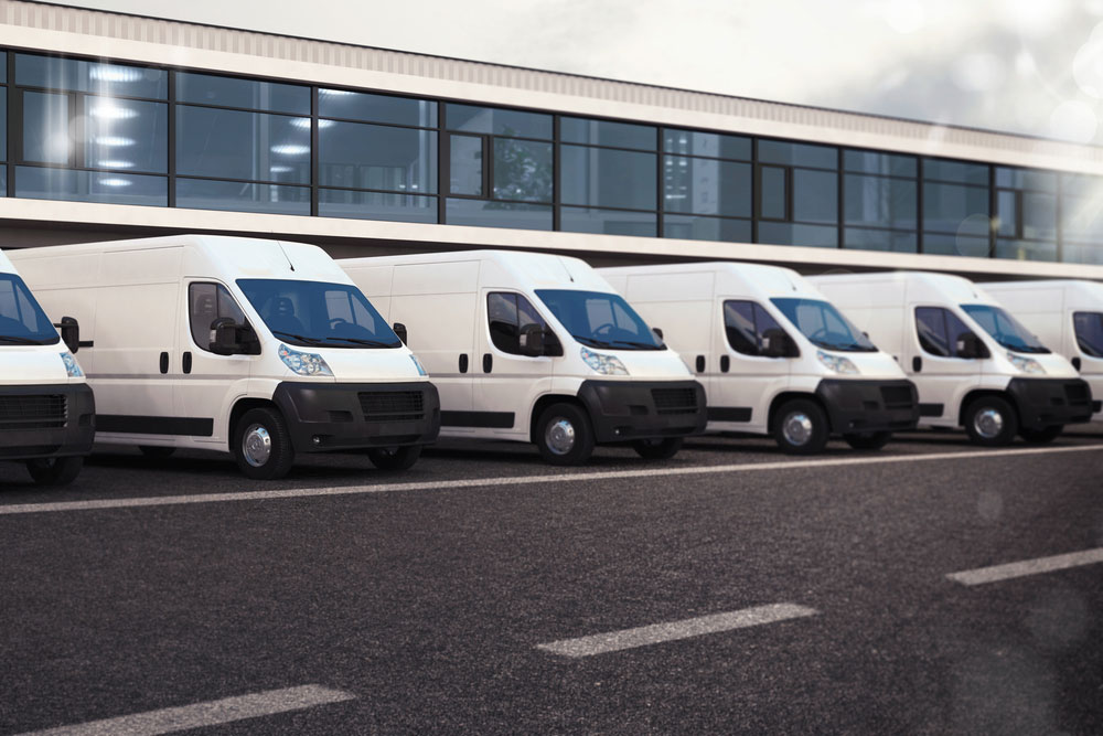 Row of white commercial vans parked in line outside modern warehouse building.