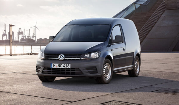 White and dark grey Volkswagen Caddy van parked on industrial estate with refinery visible in background.