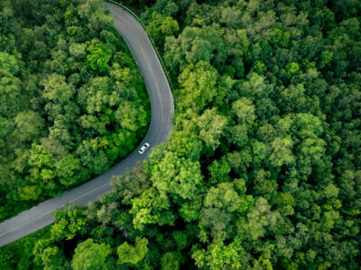 Aerial top view of electric vehicle driving on highway road in green forest. Sustainable transportation, zero emissions, and eco-friendly mobility for reducing carbon footprints. Road infrastructure.