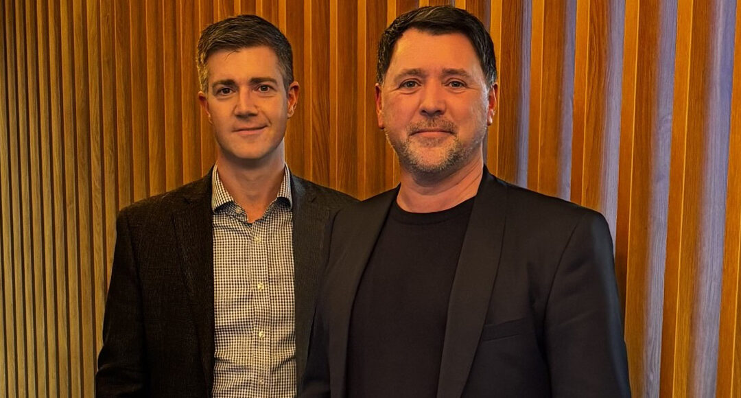Two men stand together against a wooden slatted background, one wearing a checked shirt and navy blazer, the other in a…