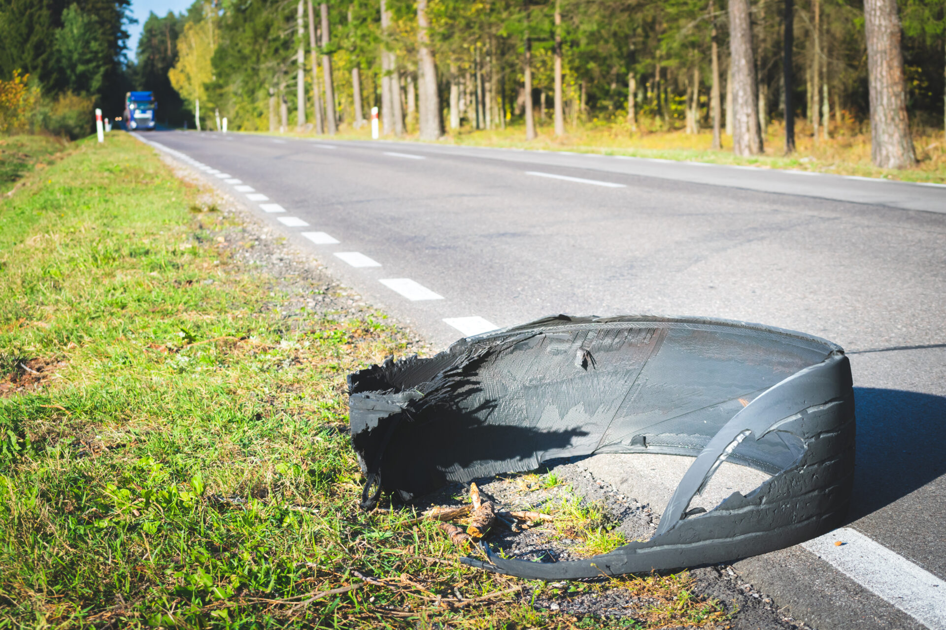 Damaged car tyre in roadside grass verge with forest and road in background.
