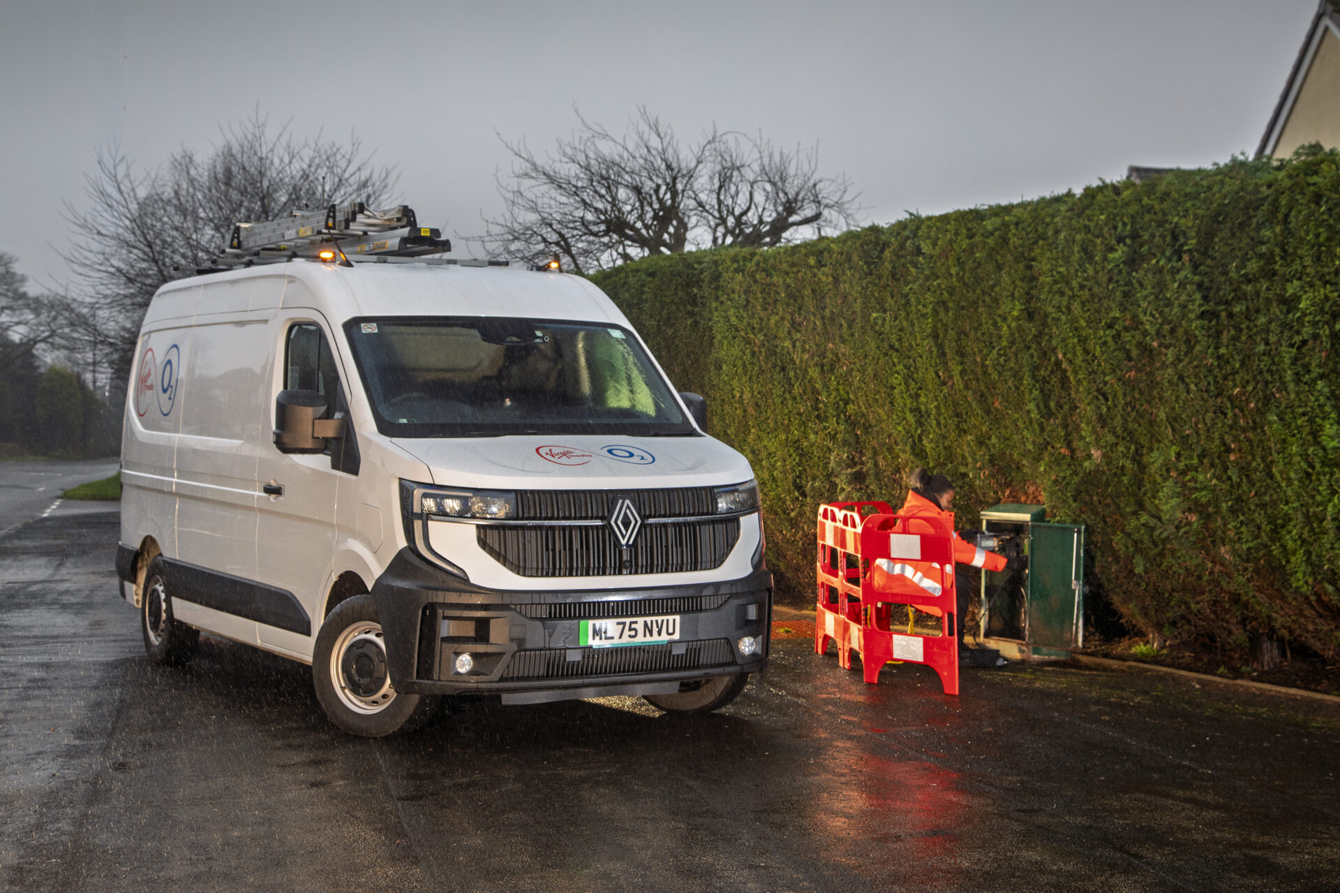 White Renault electric van with Virgin Media O2 branding parked beside red equipment on wet driveway, with evergreen hedge…