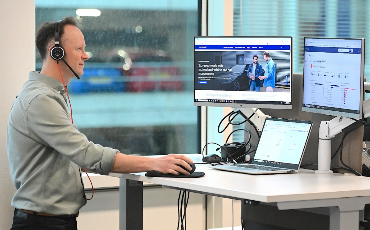 Man wearing headset at desk with dual monitors displaying fleet management software and business webpage.