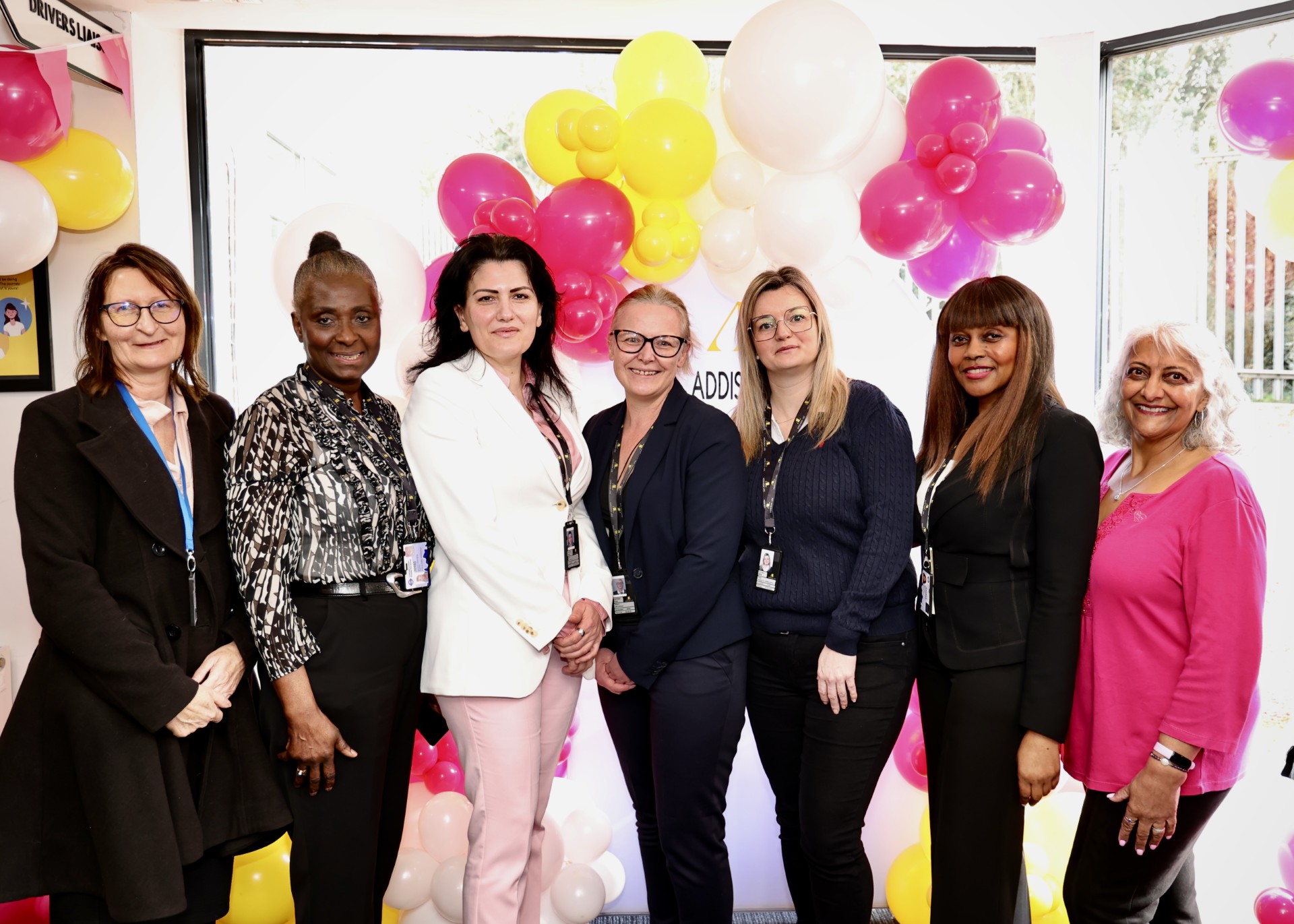 Seven women stand together in front of a backdrop decorated with pink, yellow and white balloons at a corporate event.