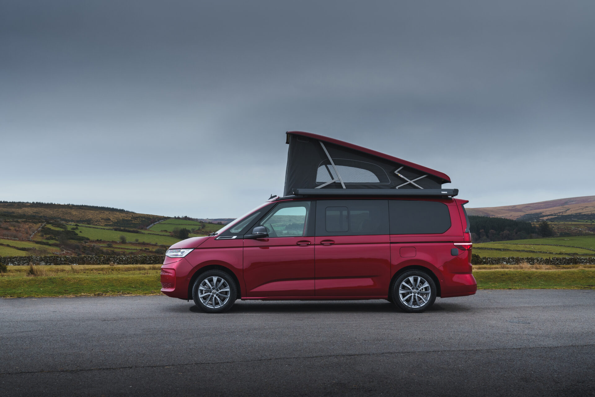 Red Volkswagen Multivan campervan with pop-up roof open, parked on road with rolling countryside hills behind.