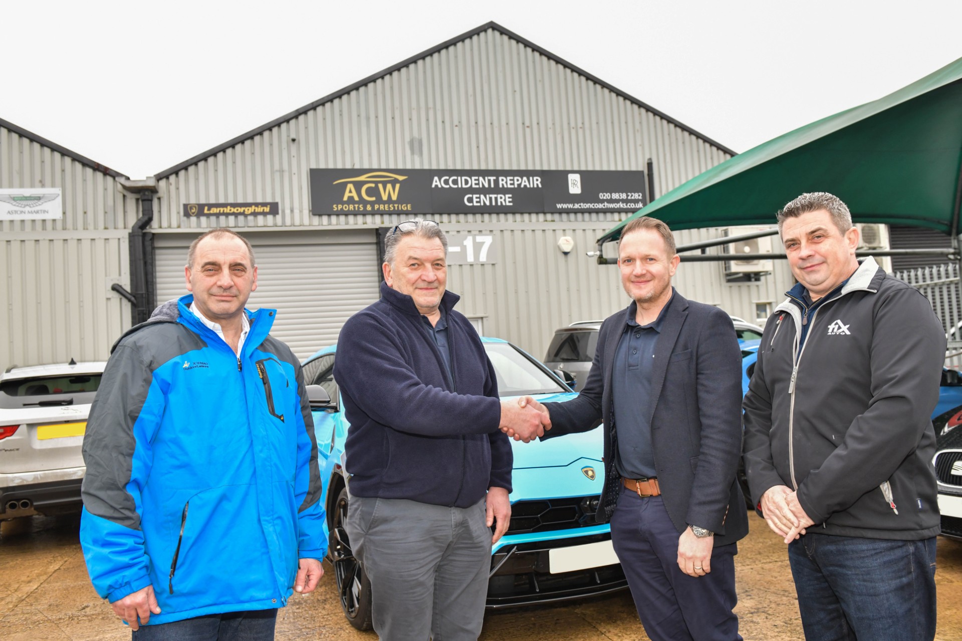 Four men shake hands outside ACW Accident Repair Centre, with vehicles and industrial buildings behind them.