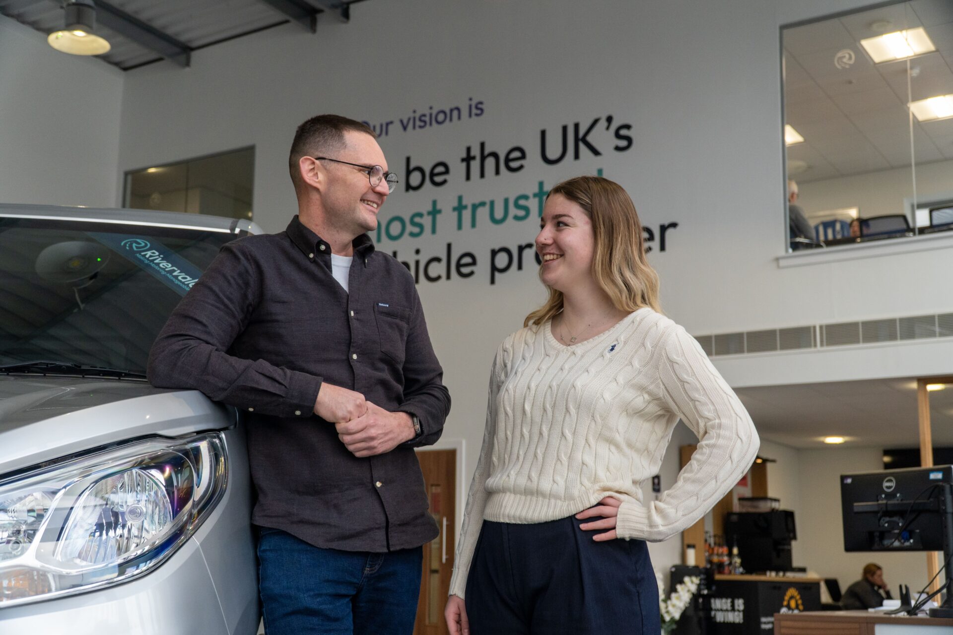Man and woman shaking hands beside a white van in a car dealership showroom with vision statement on wall behind them.