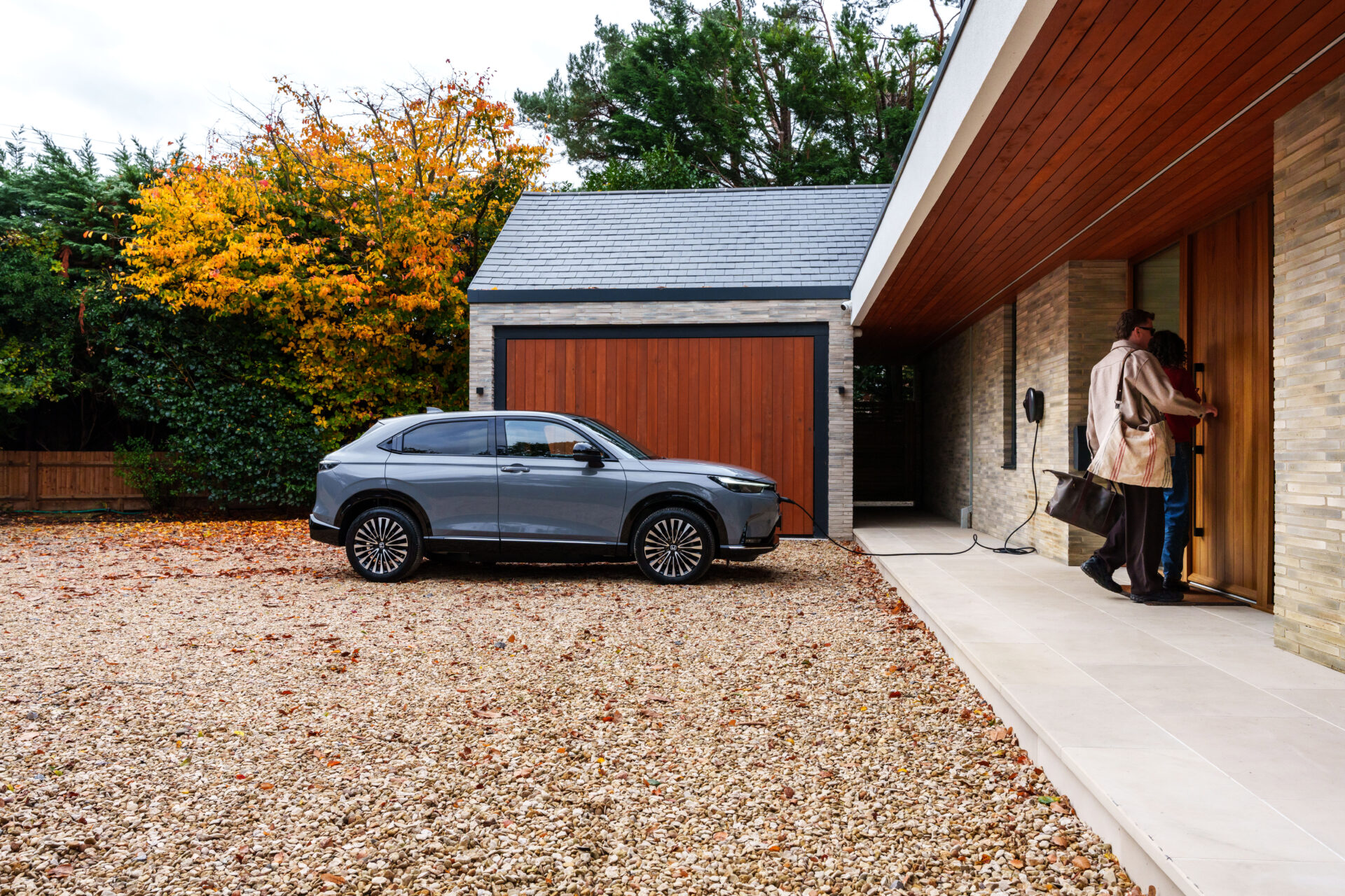 Blue electric SUV parked on gravel driveway outside modern house with red garage and wooden entrance door.