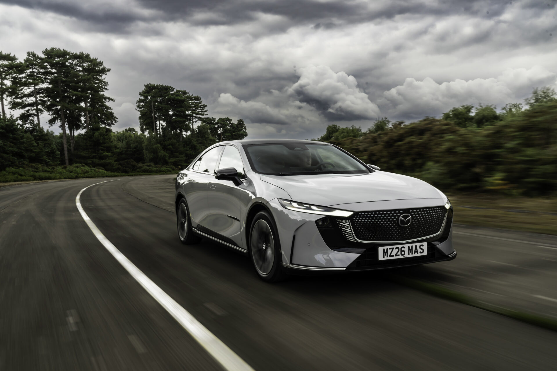 White Mazda 6e electric saloon driving on curved road with trees and dramatic sky overhead.