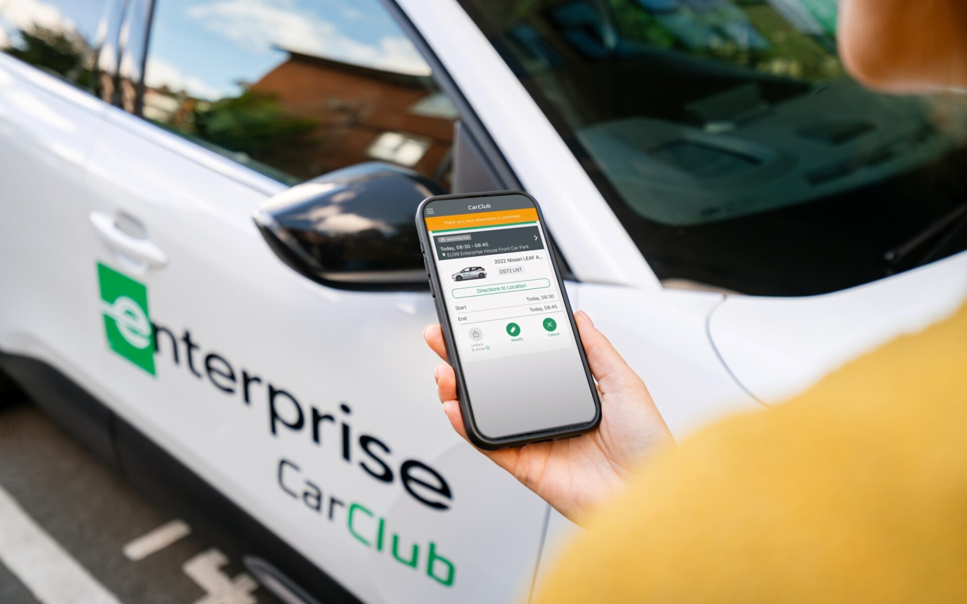 Hand holding smartphone displaying Enterprise Car Club app, with white Enterprise car club vehicle in background.