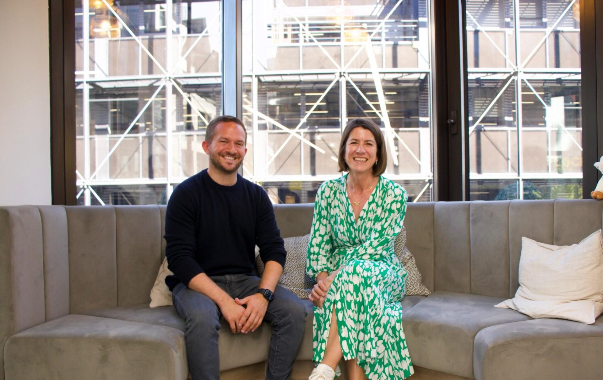 Two people sit on a beige sofa in front of large windows with geometric patterns, urban buildings visible outside.