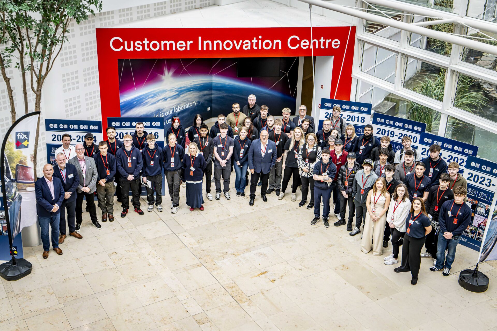 Large group of apprentices and staff members posing in front of Customer Innovation Centre display with Earth imagery and…