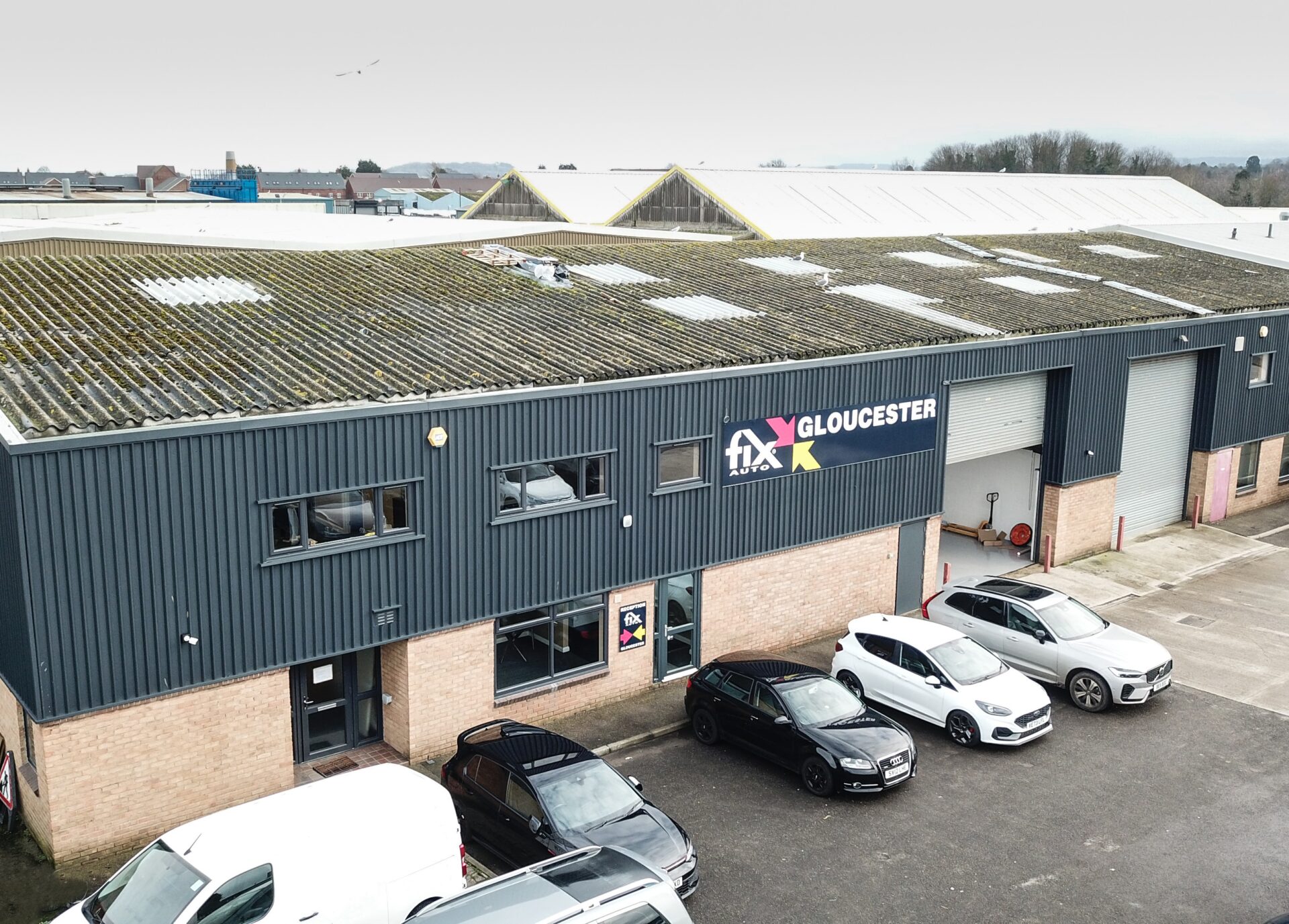 Fix Auto Gloucester facility with parked vehicles outside brick and metal-clad building, green-roofed structures behind.