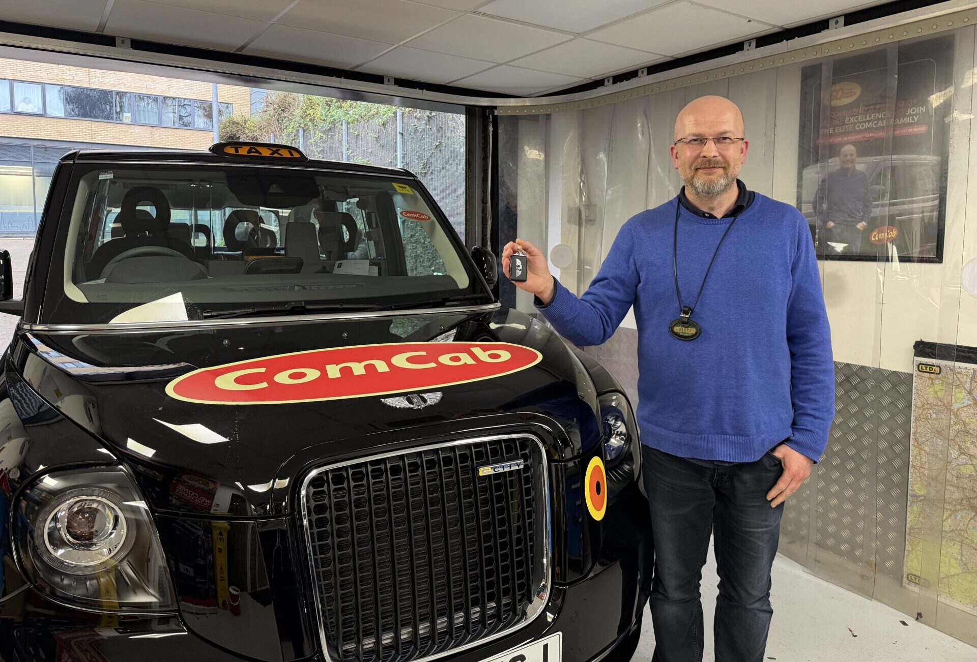 Man in blue jumper standing beside black London taxi cab with Comcab branding inside training facility.