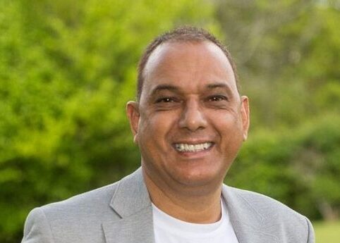 Man in grey blazer smiling at camera with green foliage background.