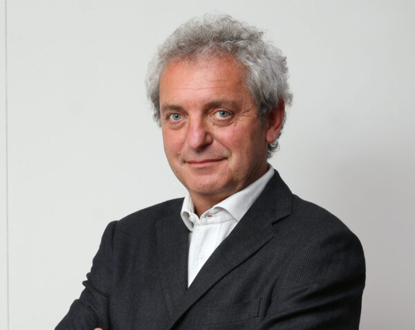 Man with grey hair in dark blazer and white shirt against white background.