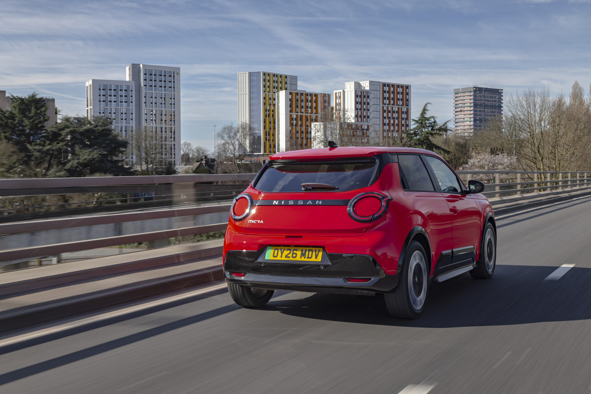 Red Nissan Micra electric car driving on urban bridge with riverside apartments in background.