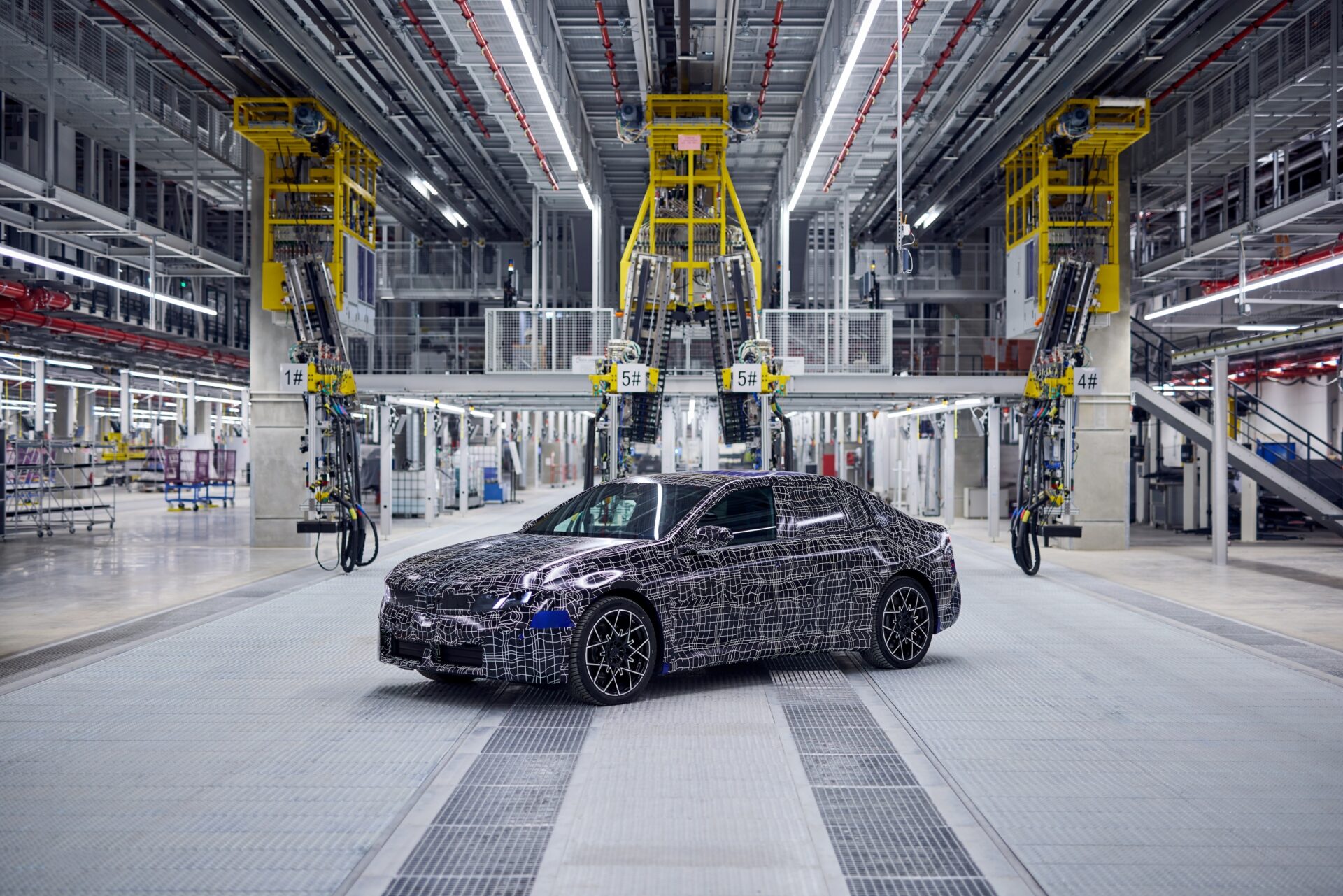 BMW electric 3 Series prototype in camouflage on assembly line surrounded by yellow robotic arms in factory.