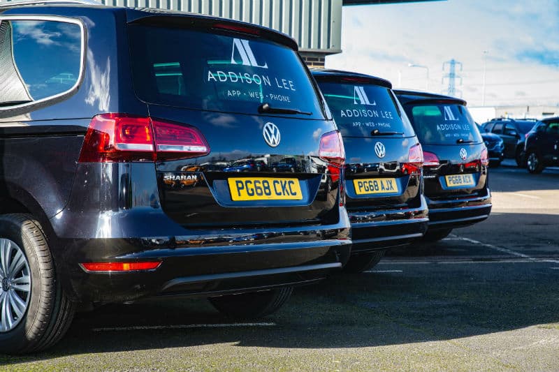 Row of blue Volkswagen Sharan taxis with Addison Lee branding parked at a depot, displaying registration plates.