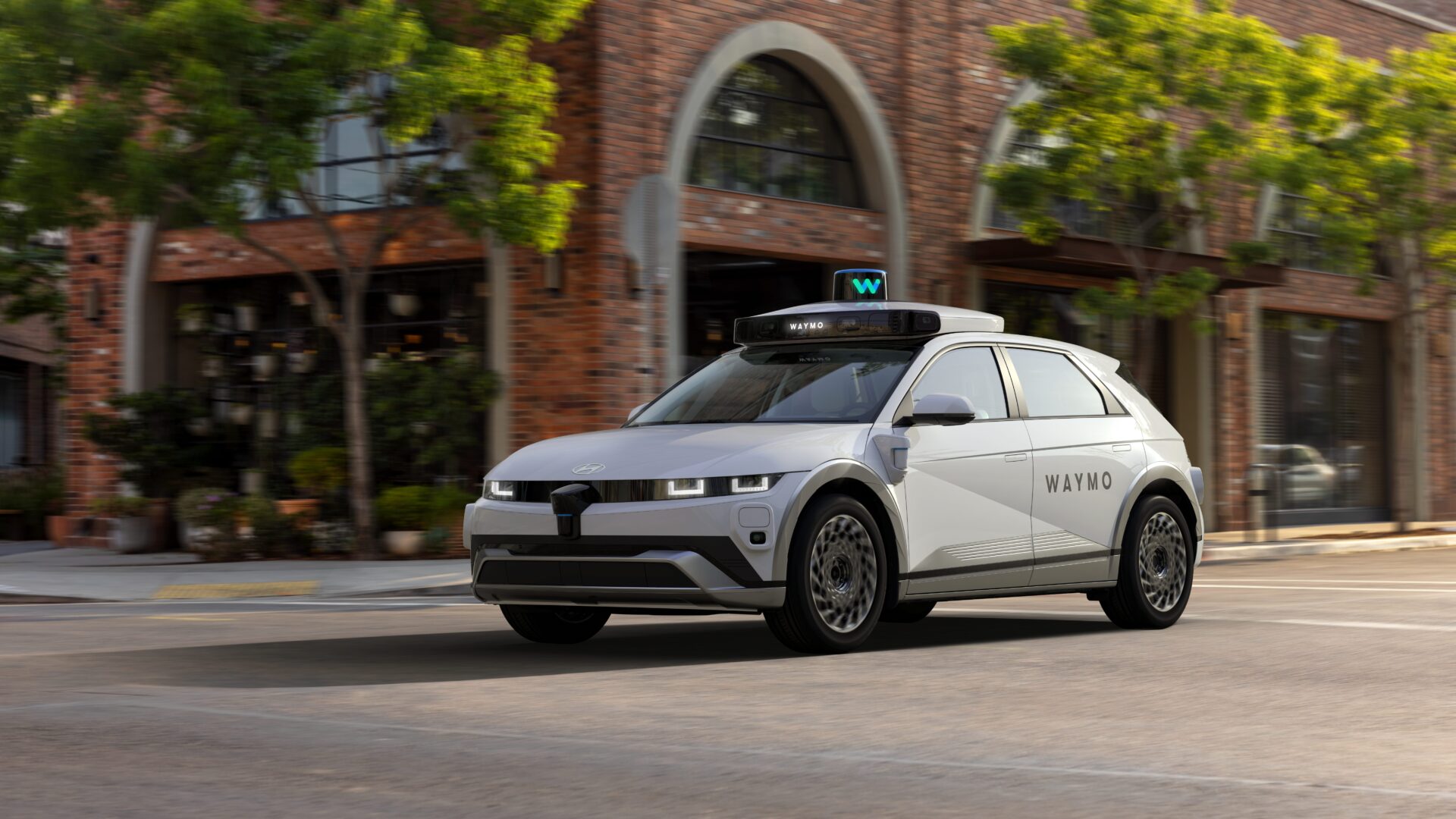 White autonomous taxi vehicle with Waymo branding parked on urban street outside brick building with ivy.