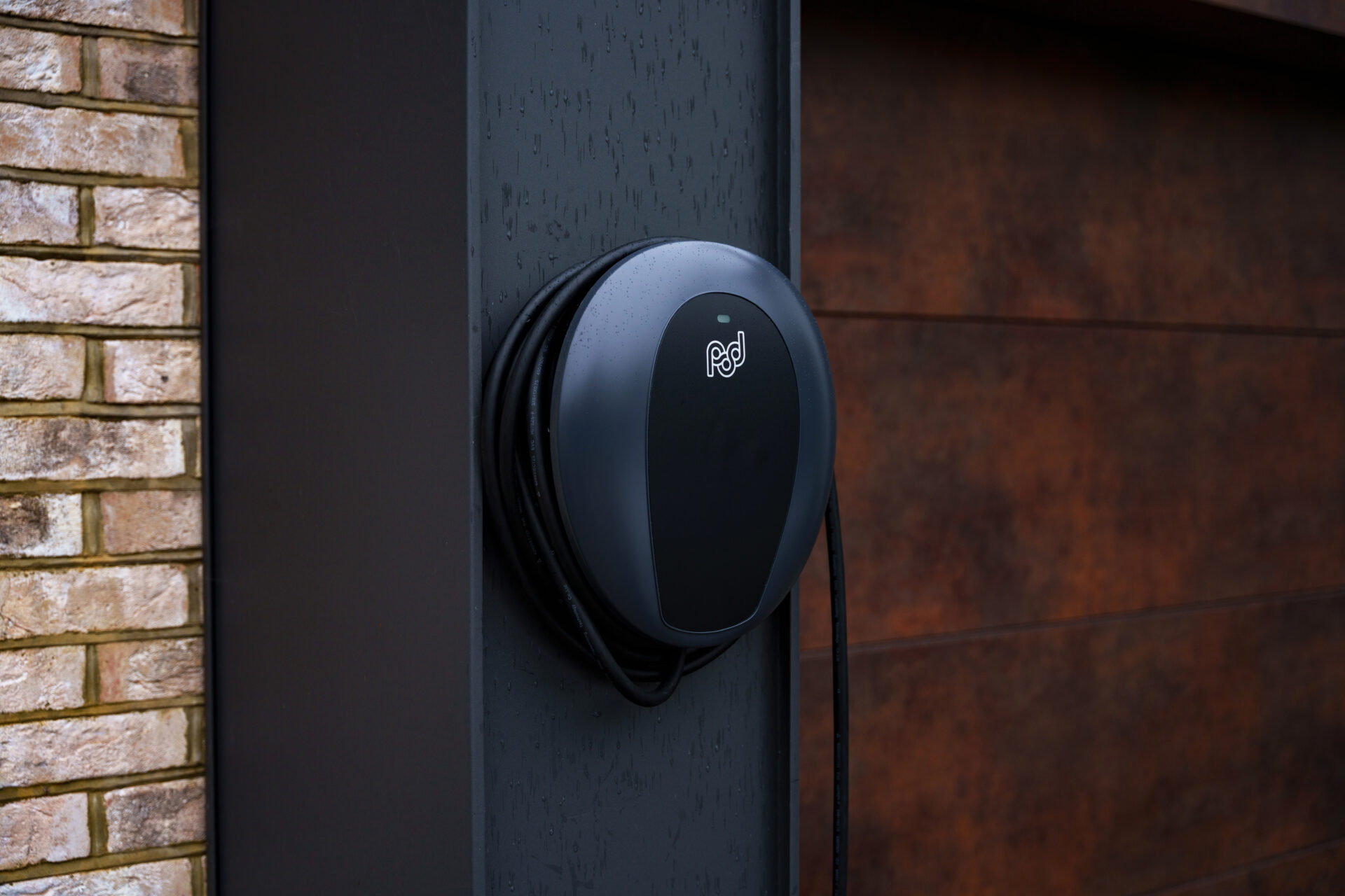 Black wall-mounted electric vehicle charging pod attached to dark metal pillar beside brick building.